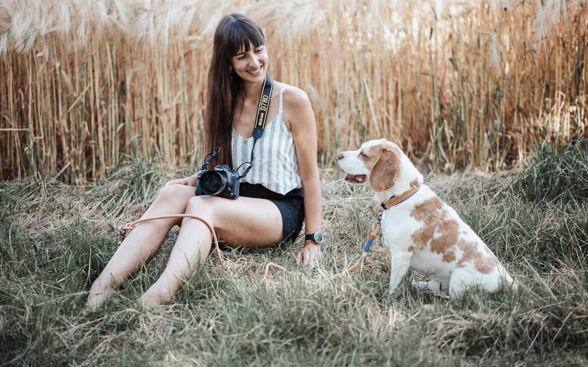 Frau sitzt in der Natur mit Kamera und lacht Hund an